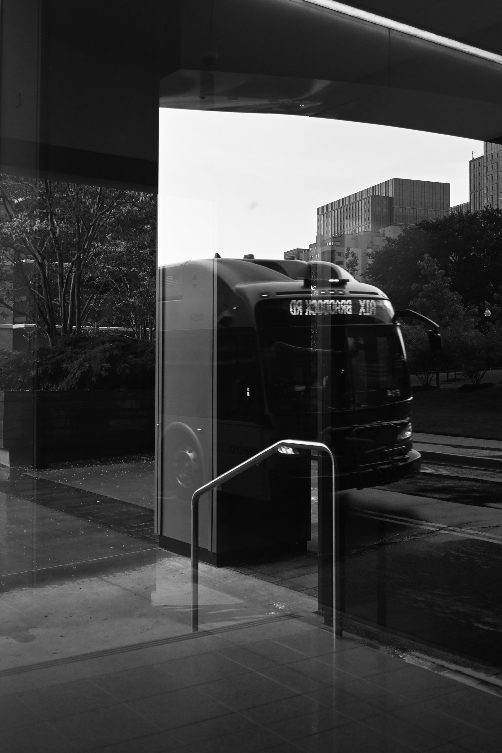 Black and white photo of a city bus reflected in a glass storefront beside a metal handrail, photographed by Mike Loerop