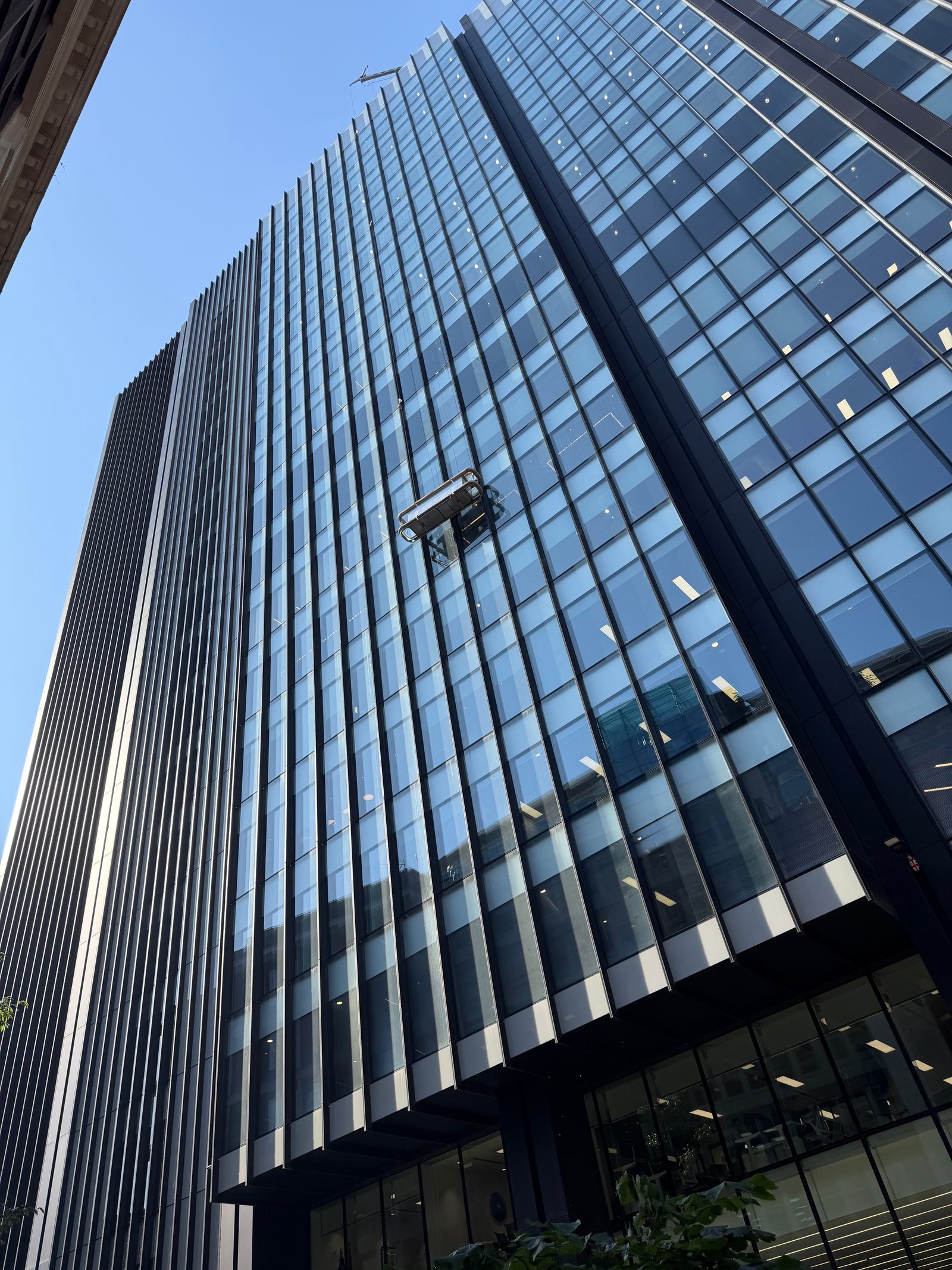 A window cleaning cradle hanging on a glass office tower in the City of London, photographed from below by Mike Loerop