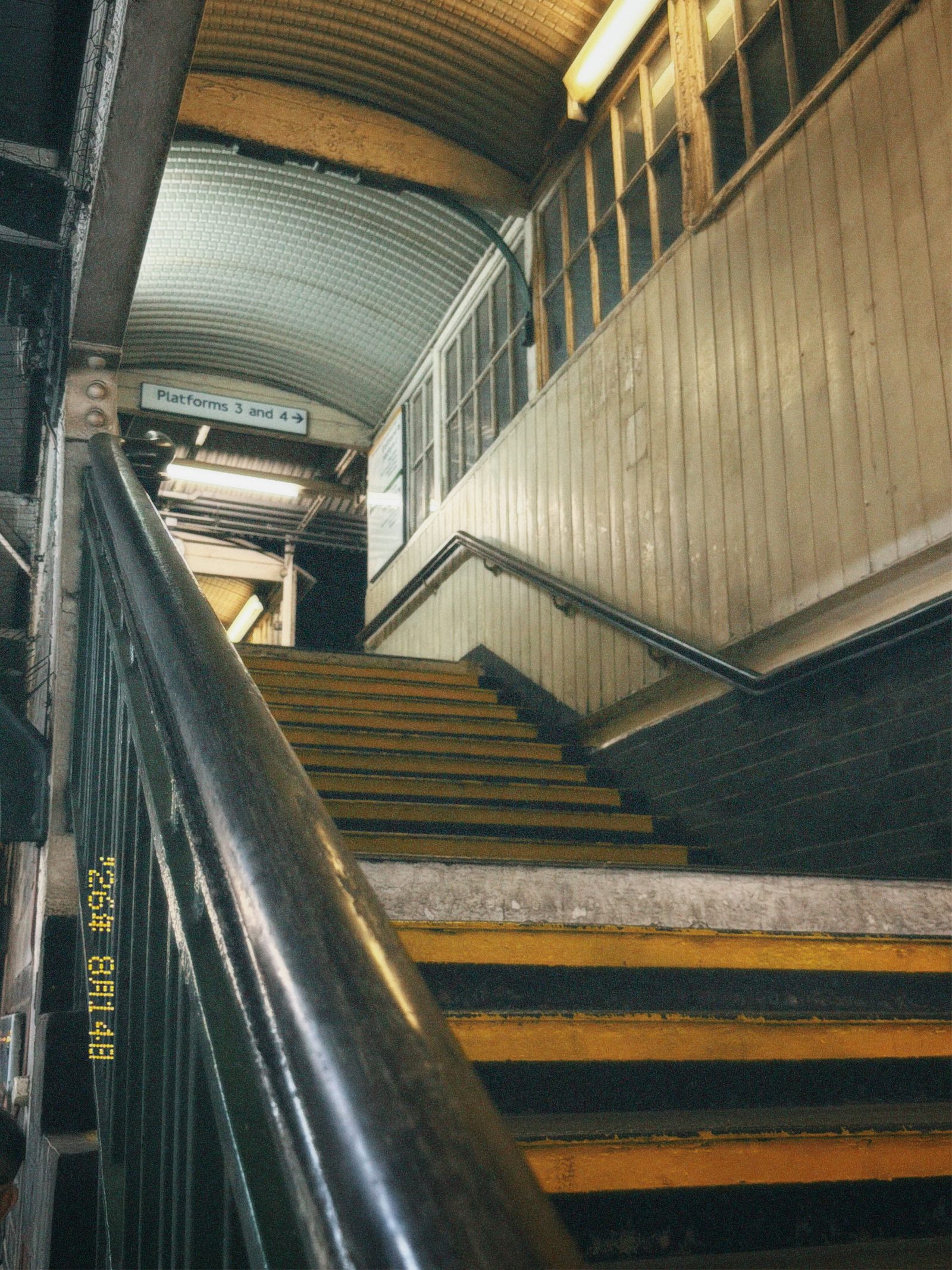 Yellow-edged station stairs and a curved metal handrail beneath a sign for platforms 3 and 4 in London, photographed by Mike Loerop