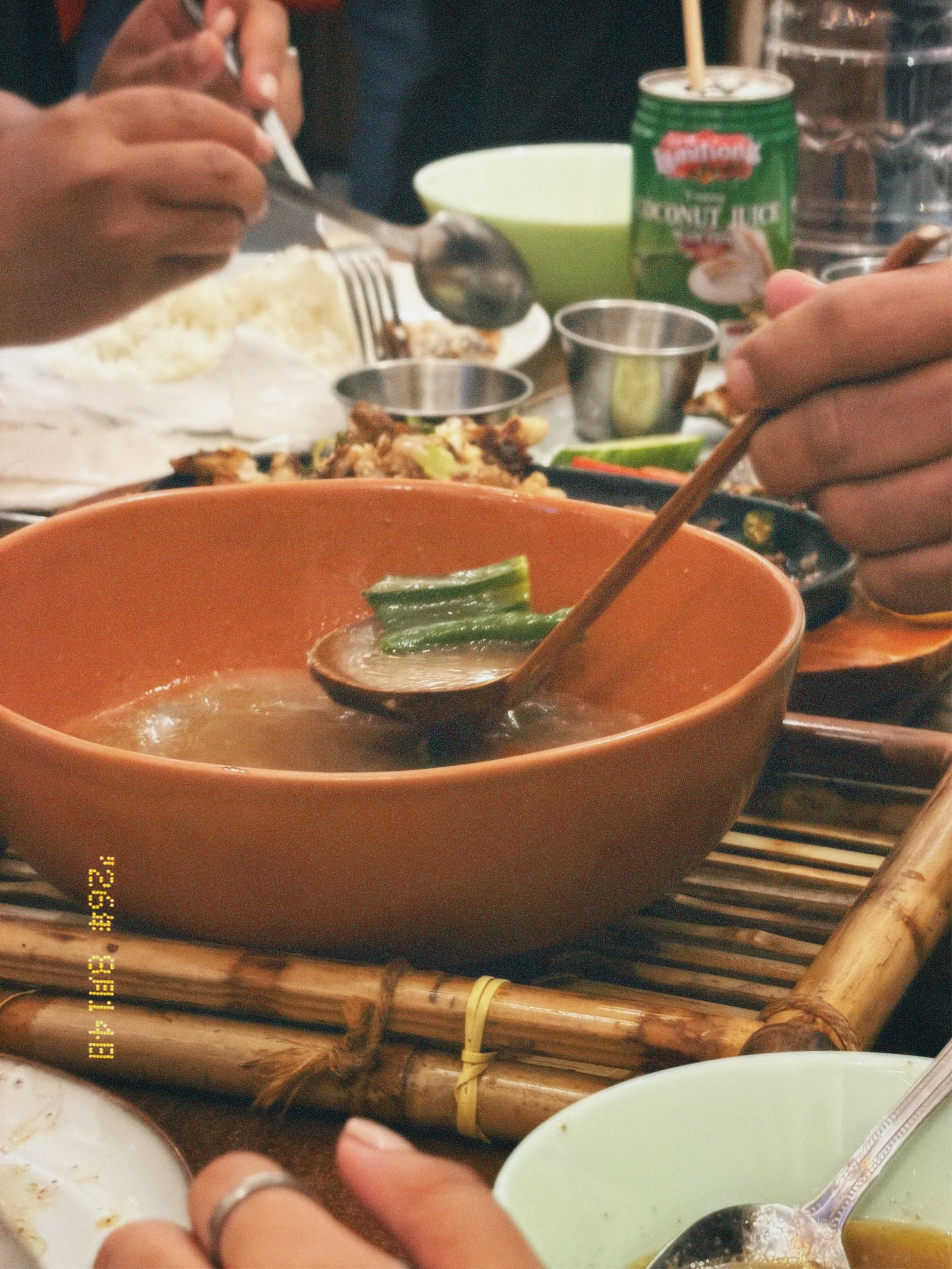 Hands reaching across a shared dinner table around a terracotta bowl of soup on a bamboo mat, photographed by Mike Loerop