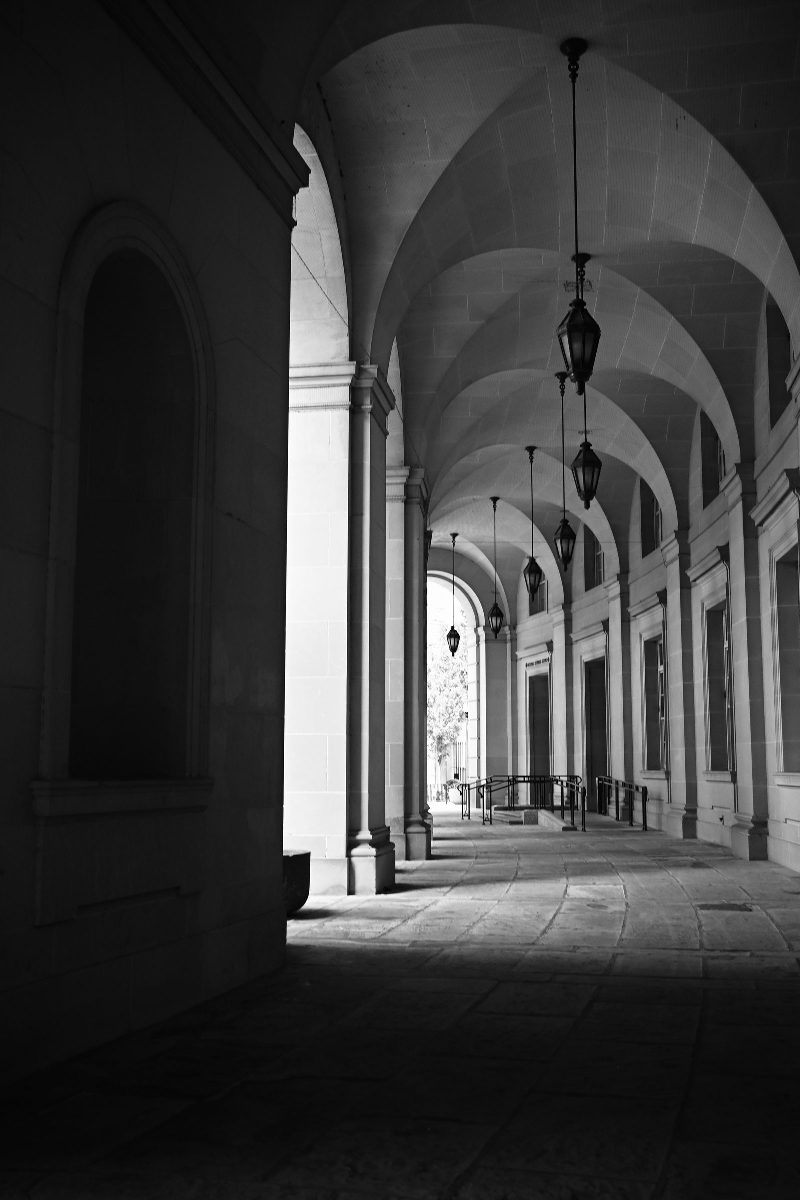 Black and white photo of a vaulted stone arcade with hanging lanterns leading toward the light, photographed by Mike Loerop