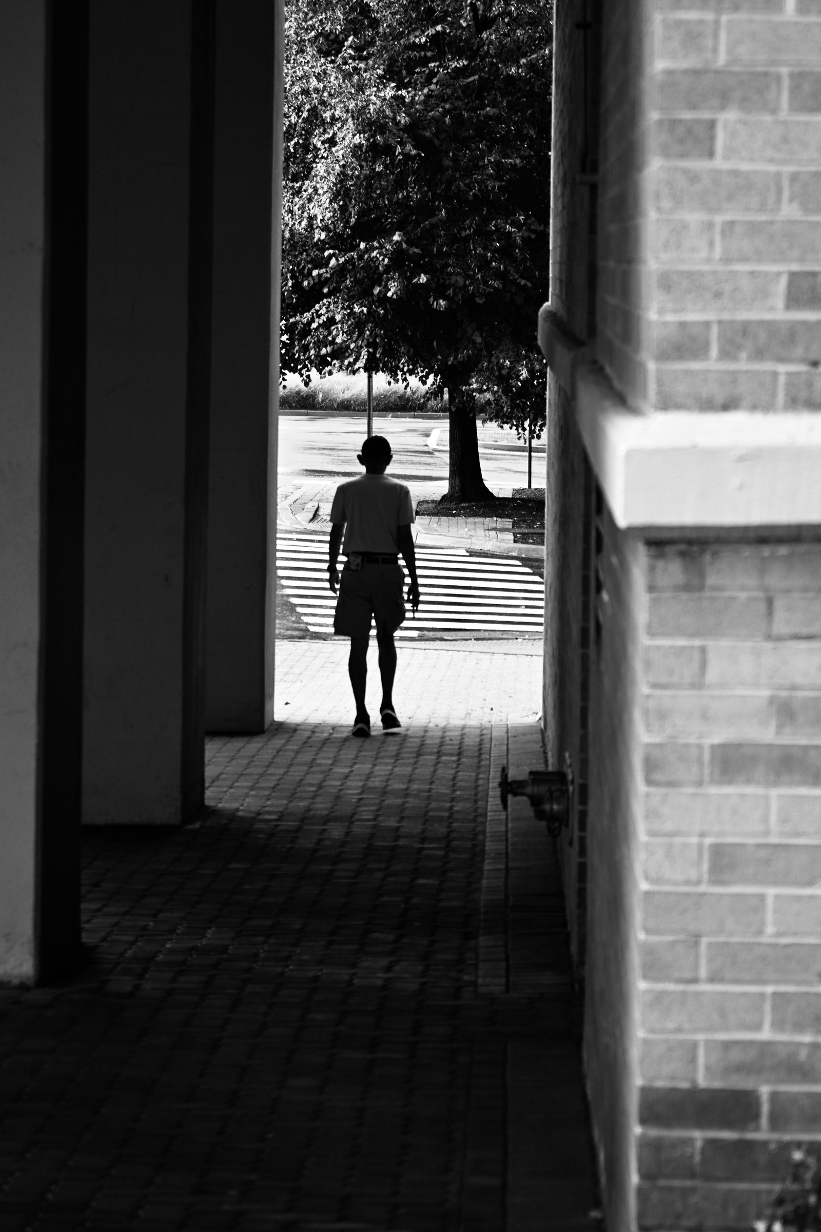 Black and white silhouette of a man walking down a shaded brick passage toward a sunlit street, photographed by Mike Loerop